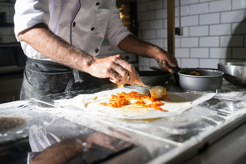 pexels-photo-8629123-8629123 Professional chef spreading tomato sauce on dough in a commercial kitchen.